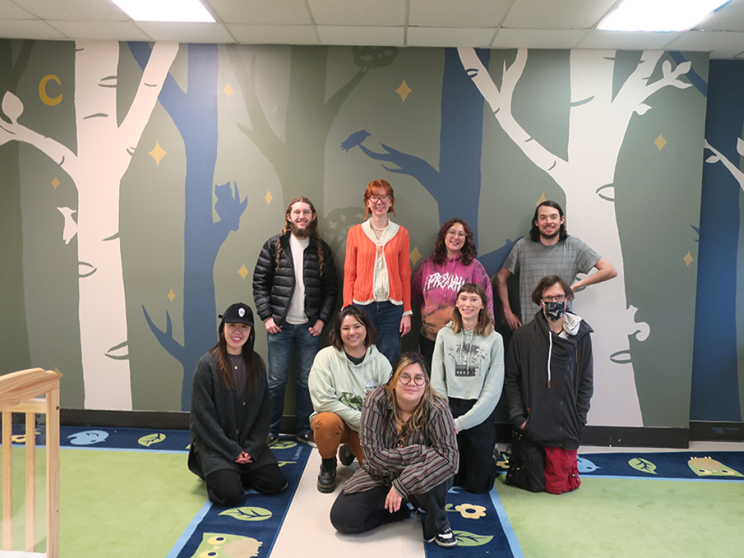 ACC Studio Art Capstone Class in front of a mural of trees and animals in ACC's Child Development Center: Tristan Dunning, Sophia Flores, Carissa Geraud, Nathan Klein, Eun Mi Lam, Lena LeBlanc, Max McMann, Jordan Stuart, and Tami Tamayo