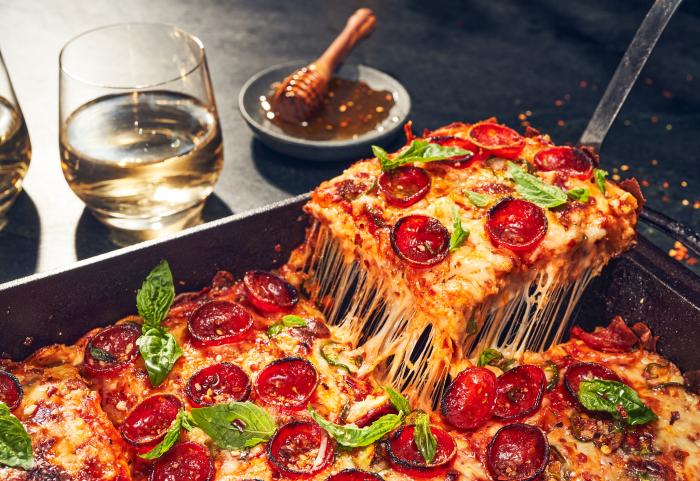 Close up of a pan of pepperoni pizza with one slice being removed. A glass of wine and bowl of honey are in the background - photo by Kate Blakeman