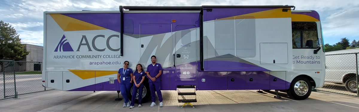 ACC's Mobile Healthcare Learning Lab with 3 EMT students standing beside it in the Littleton Campus parking lot.