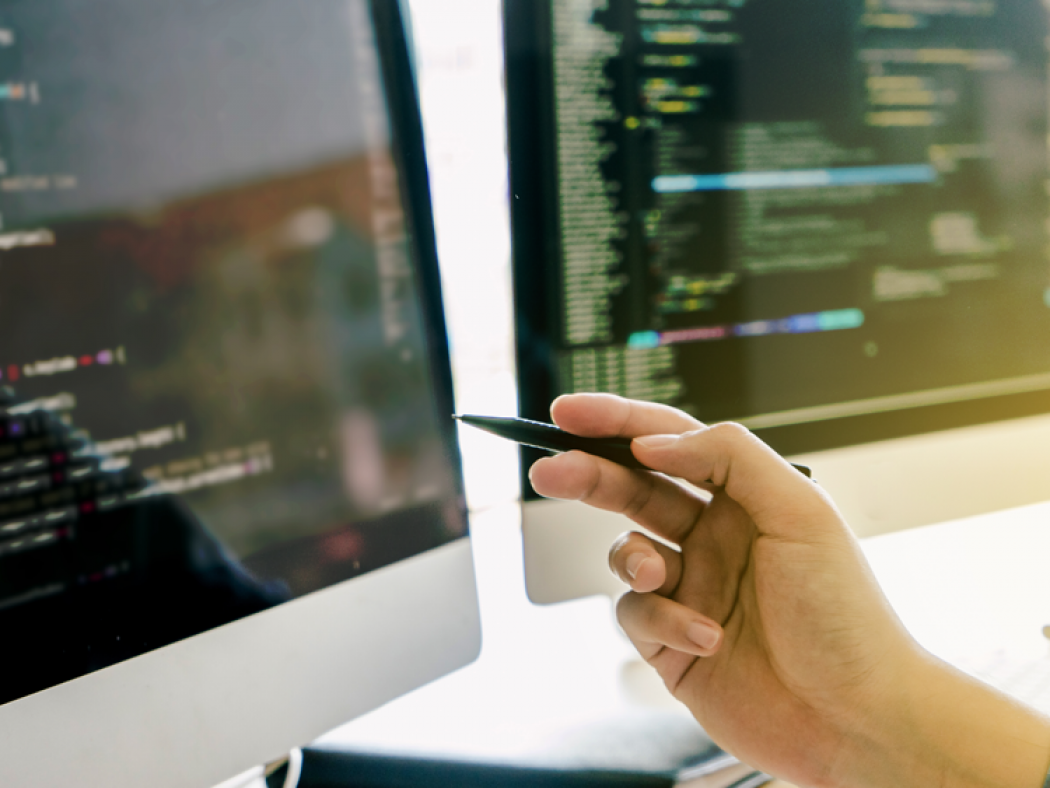 Two computer monitors with computer code on them and a hand pointing to one of the monitors.