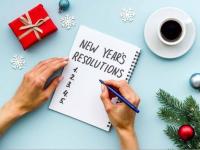 Hands writing "New Year's Resolutions" on paper with coffee and holiday decor around.